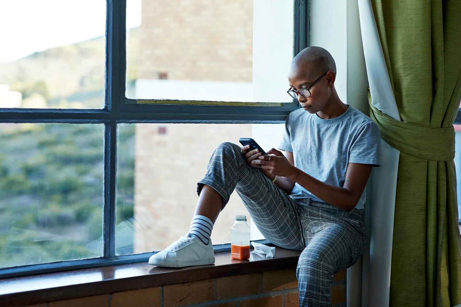 Person with glasses sitting on a windowsill looking at a smartphone, with a juice bottle nearby.