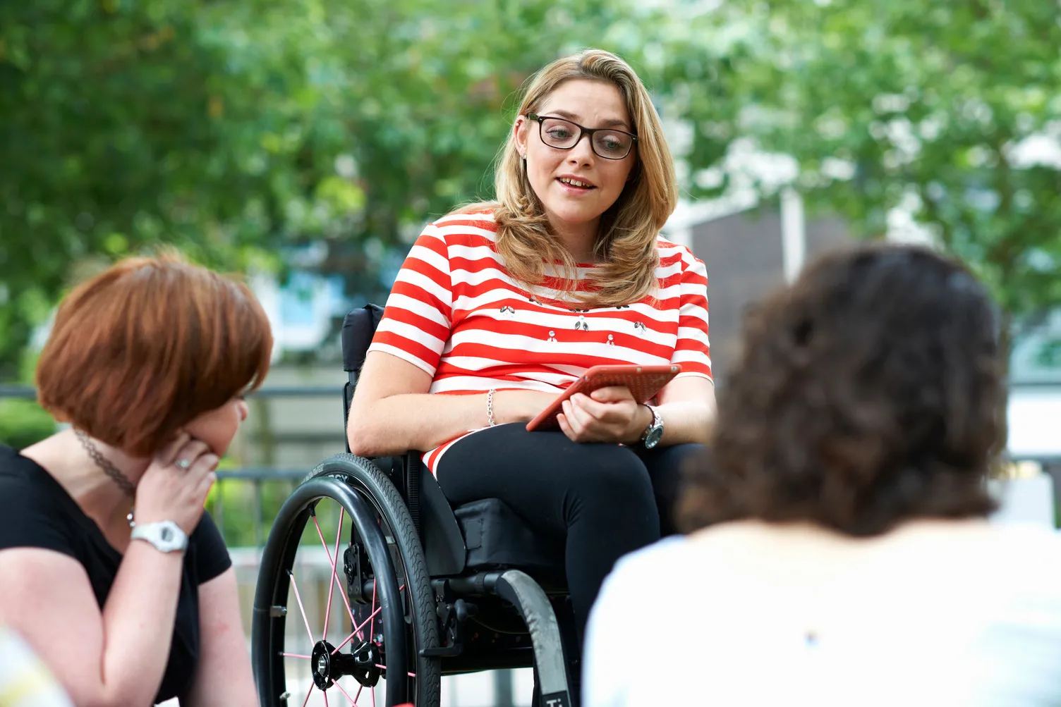 Woman in wheelchair holding a tablet, talking to two seated women outdoors with green trees behind.