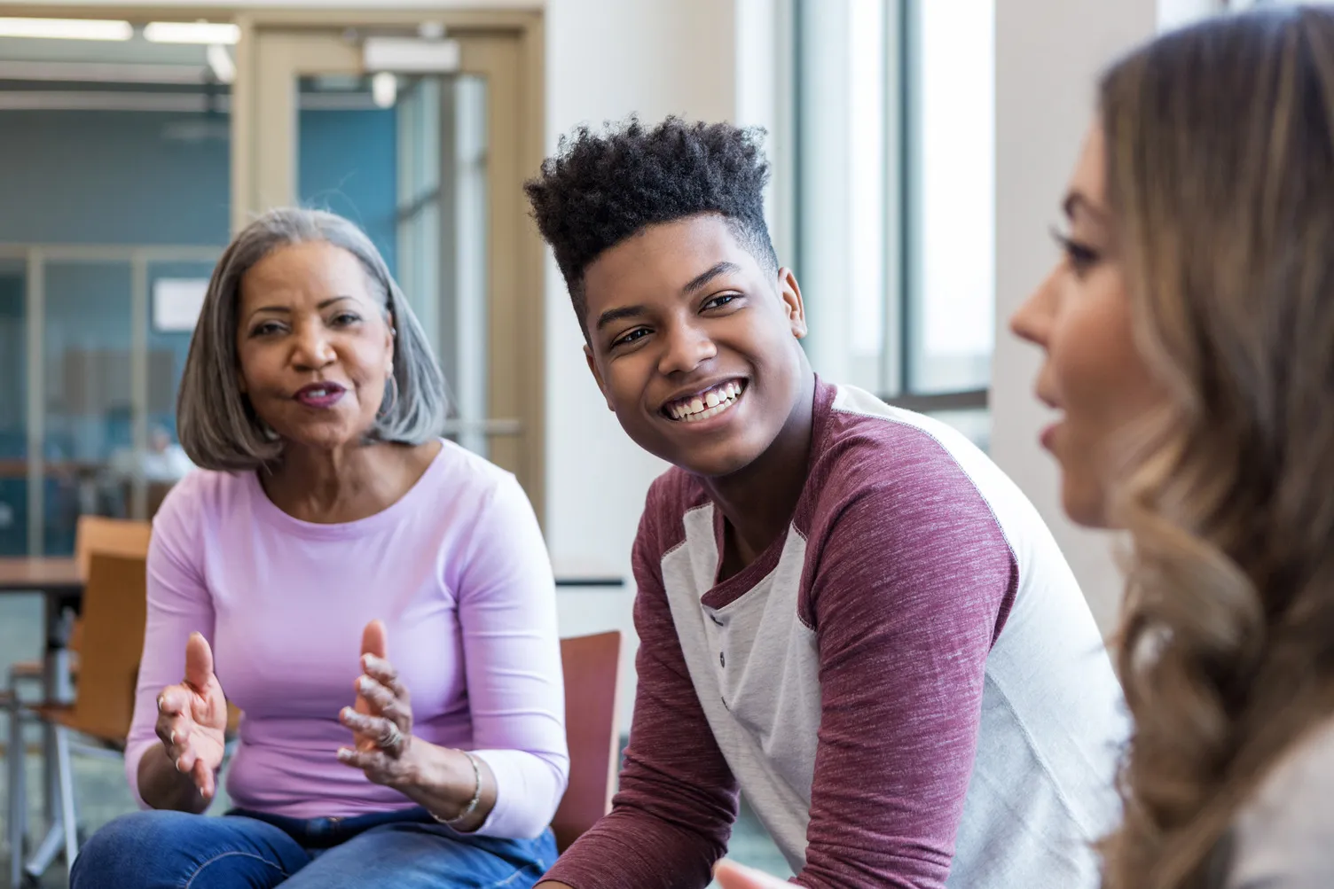 Three people engaged in a discussion indoors, one older woman gesturing and a young man smiling.