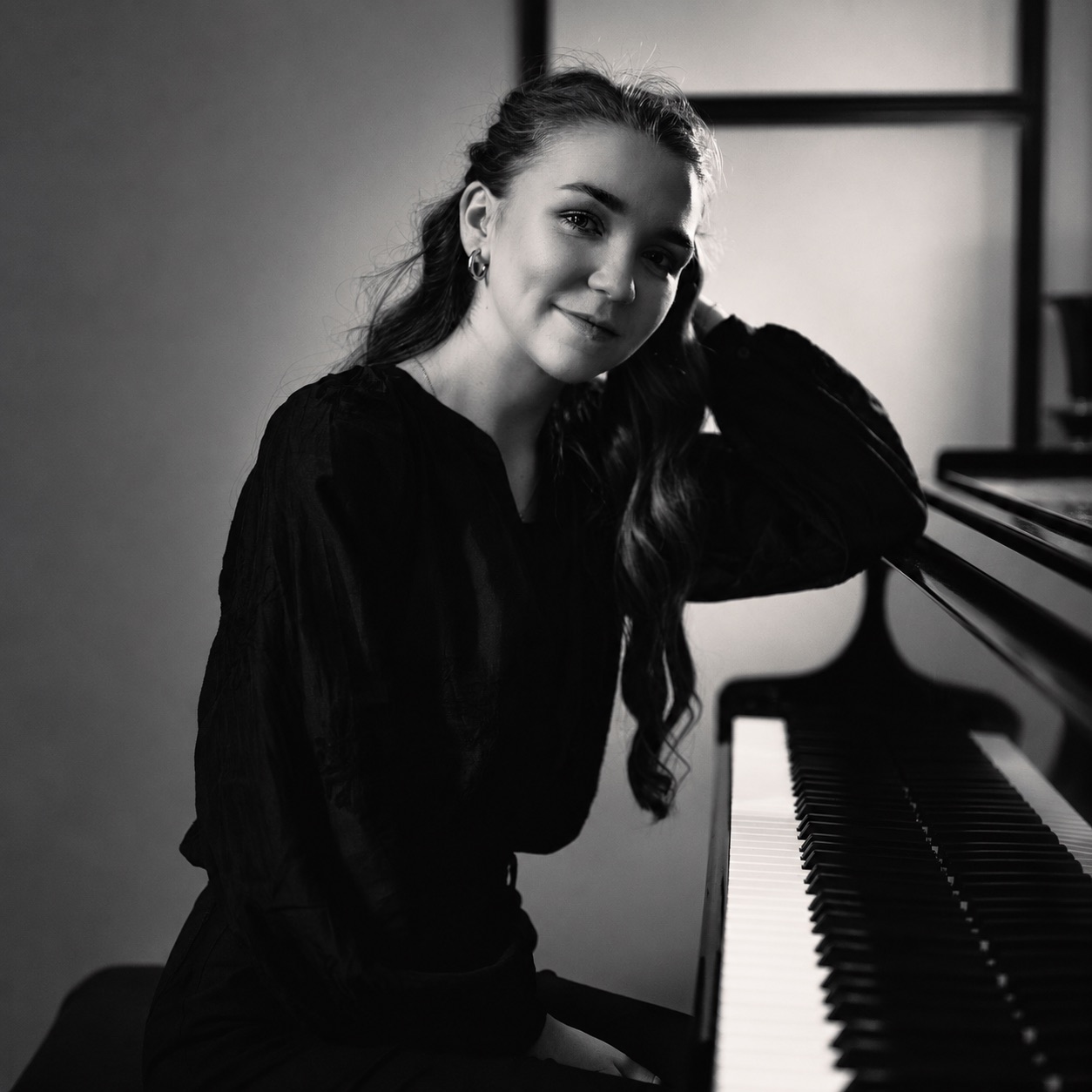Black-and-white portrait of a woman seated at a piano, smiling with her hand resting on her head.