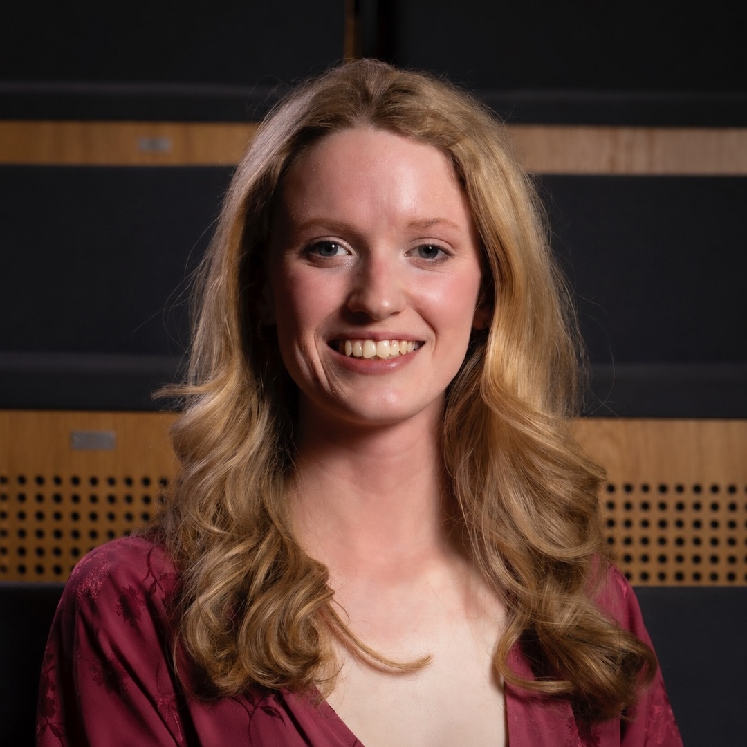 Smiling woman with long blonde hair wearing a deep red patterned blouse against dark wood background. One of the teachers.