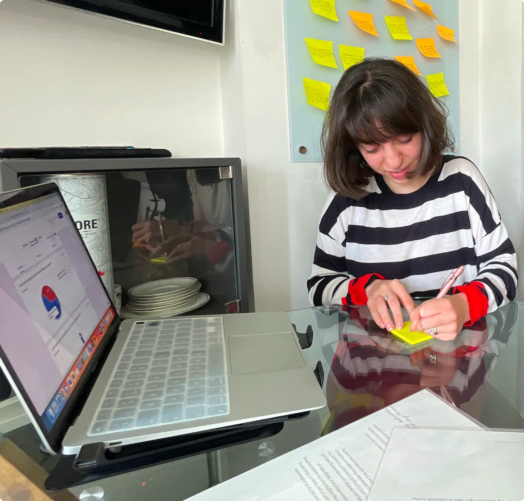 Clara Cristea, remote UX designer with a psychology background, writing notes on a yellow sticky note during a user research session at a desk with a laptop and research materials.