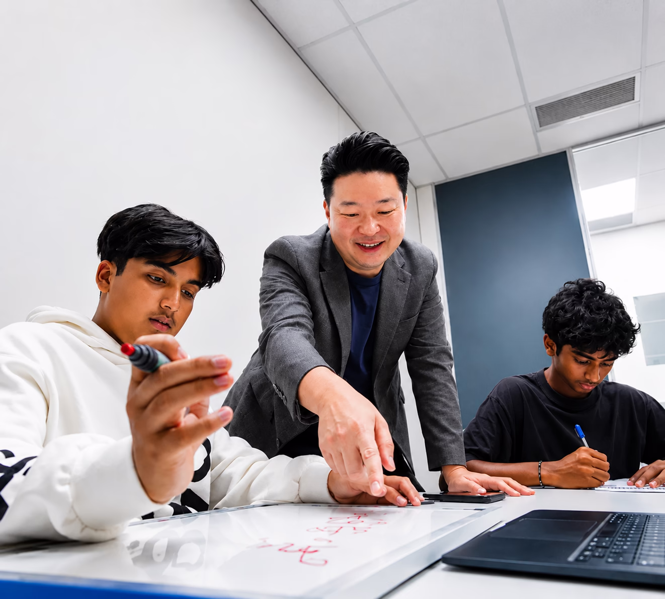 Tutor helping two students working on a whiteboard and paper in a classroom setting.