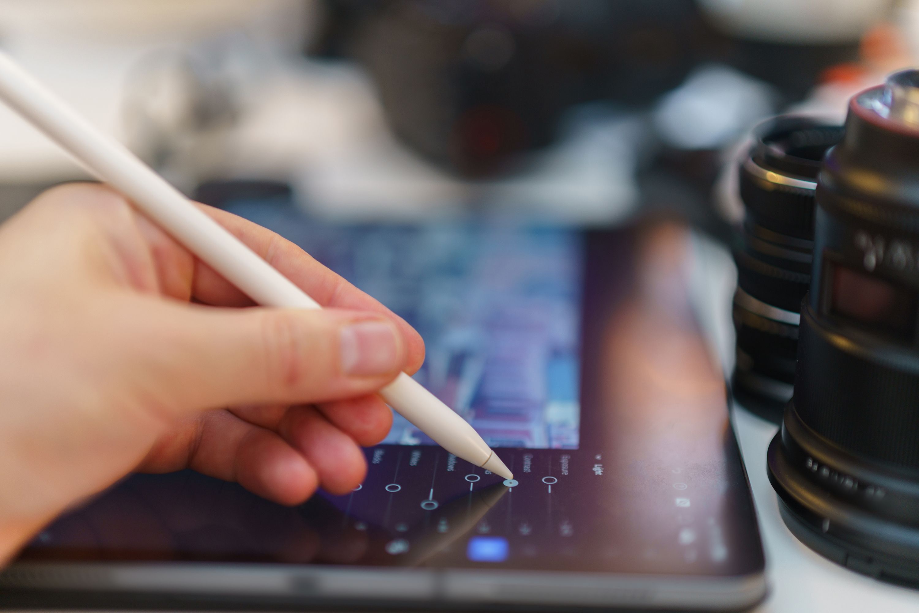 An editor adjusting photo settings on a tablet with a stylus, beside camera lenses