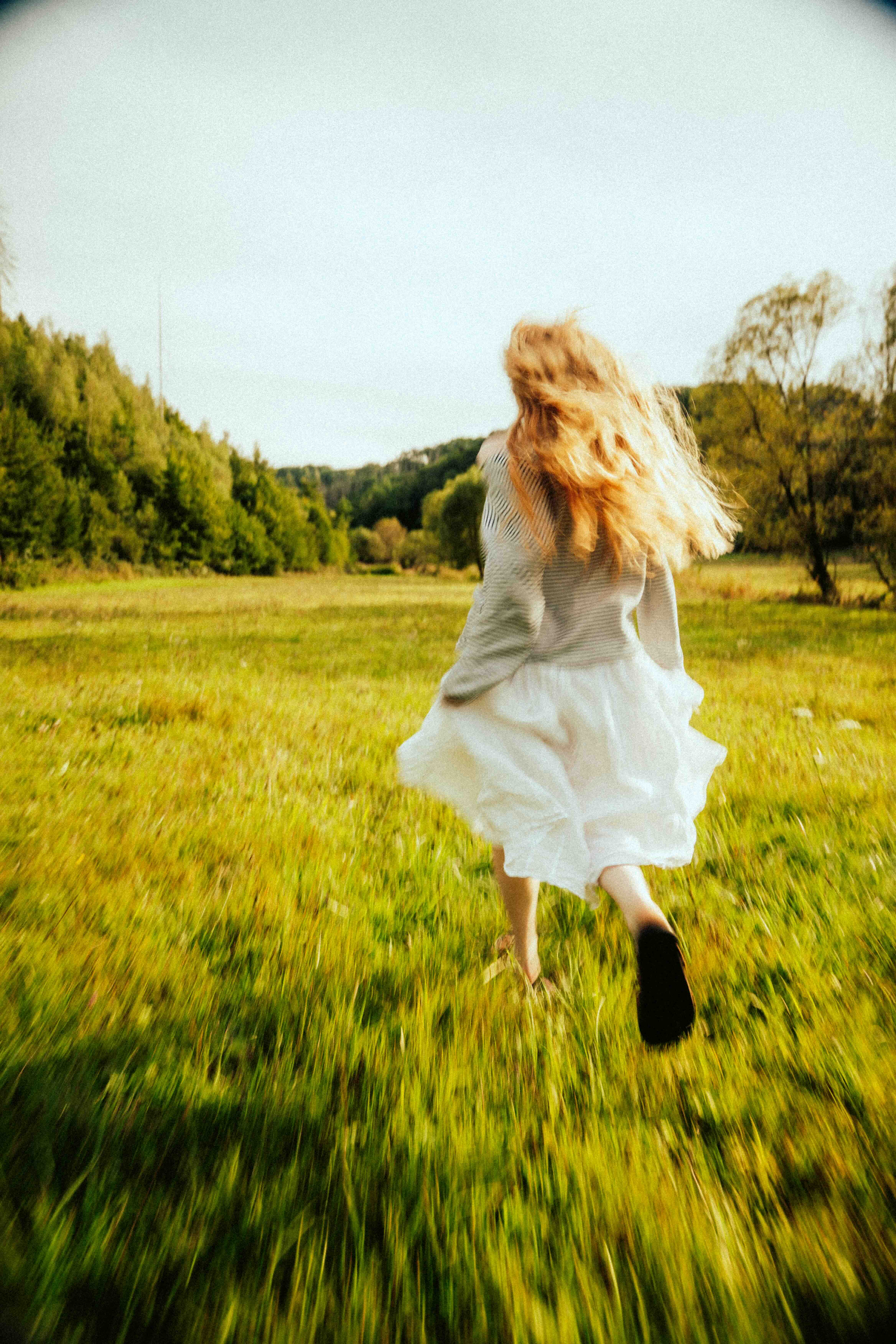 Frau im weißen Kleid läuft durch eine sonnige Wiese vor einem Waldrand