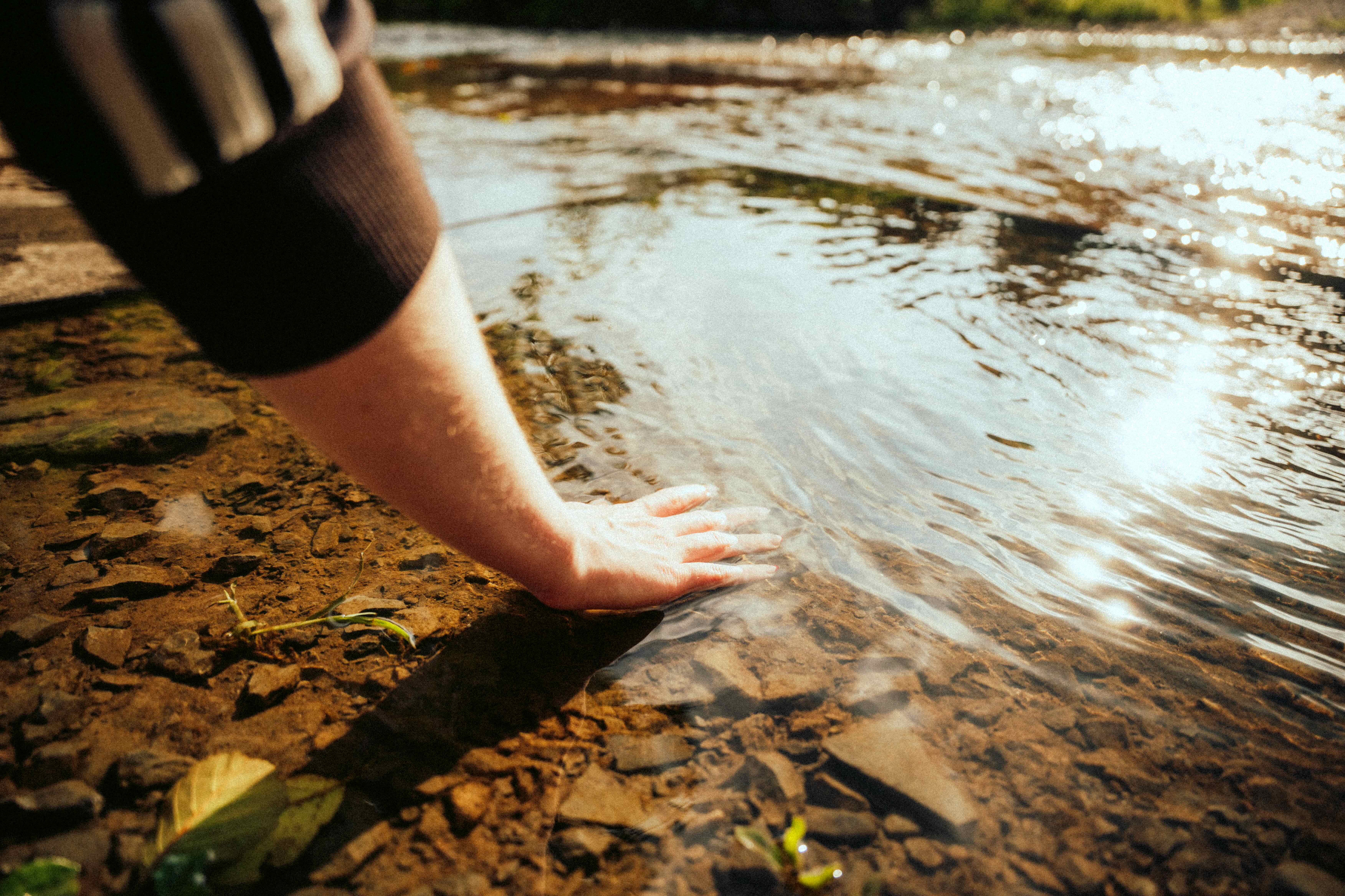 Hand berührt das flache Wasser eines Bachs über steinigem Grund. Bei Cabin Waldbach