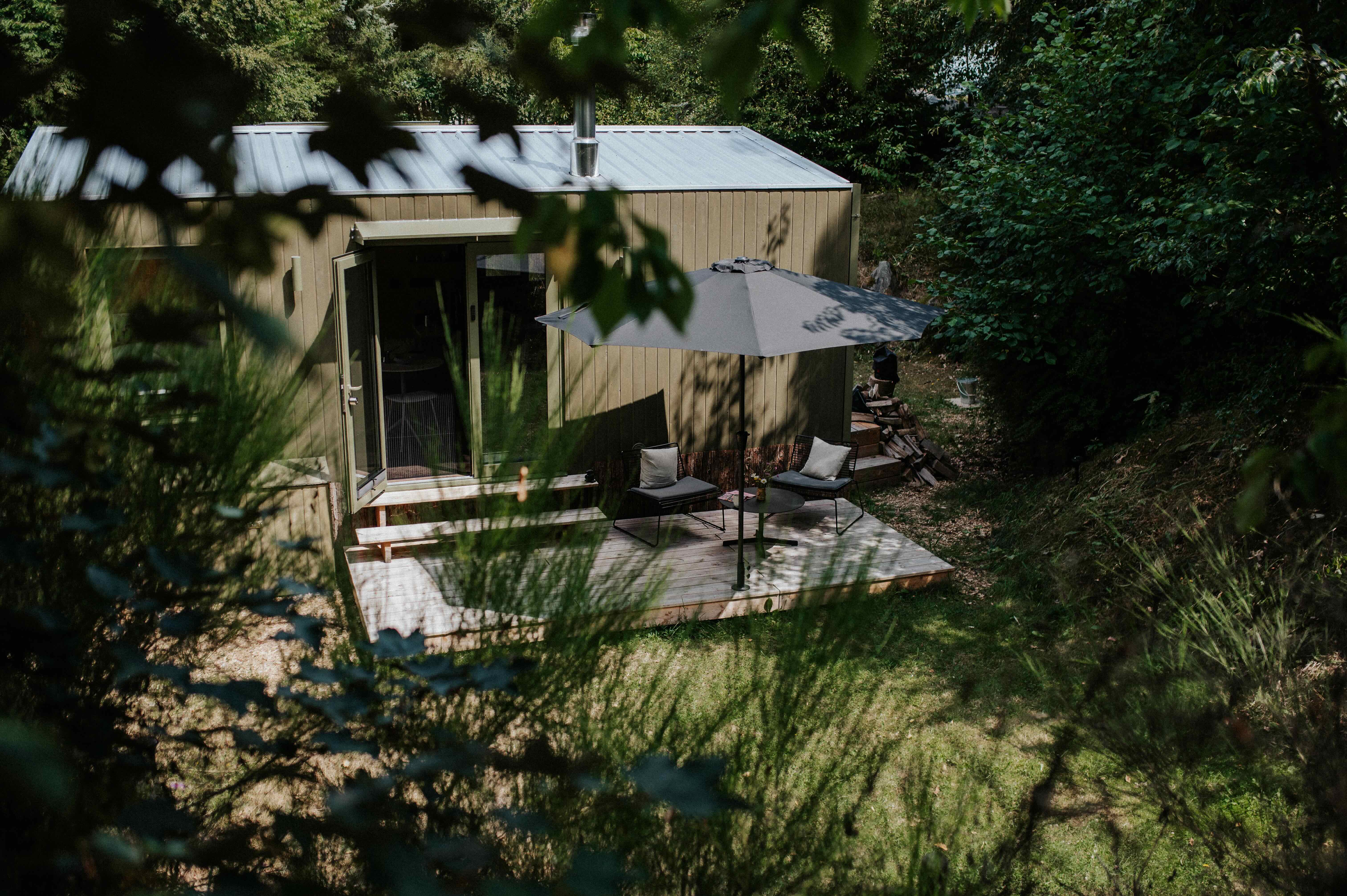 Cabin mit Holzterrasse, Sonnenschirm und Feuerholz, umgeben von dichtem Grün