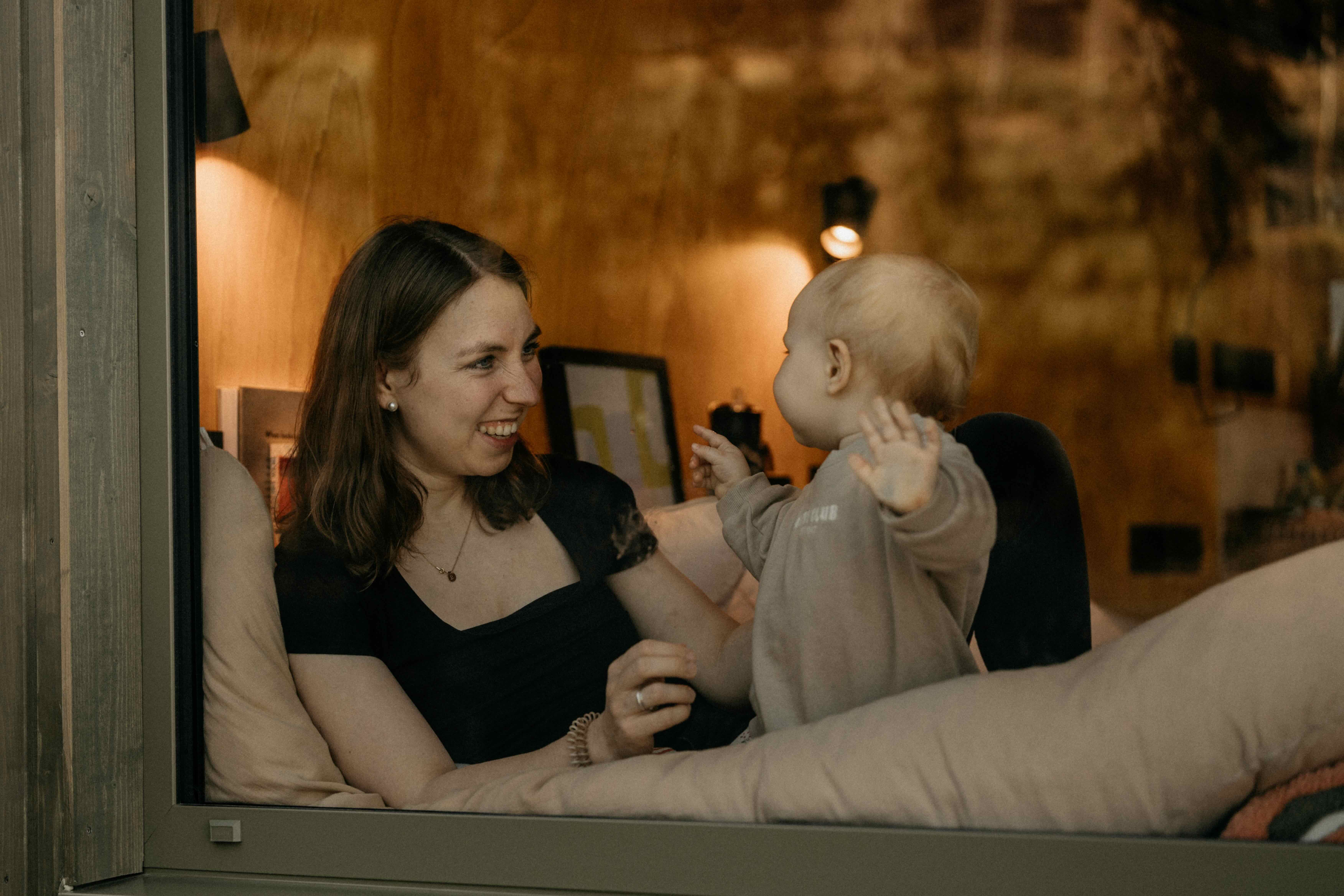Blick von außen durch das Fenster einer Cabin auf eine Frau, die mit ihrem Baby im Bett spielt