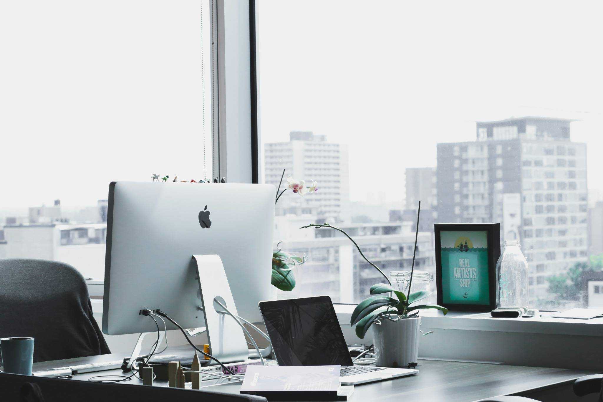 Bureau moderne – Un espace de travail lumineux avec ordinateurs Apple, carnet et plante, donnant sur une vue urbaine.