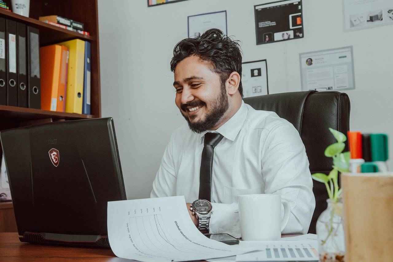 Un homme en chemise et cravate souriant devant son ordinateur au bureau.