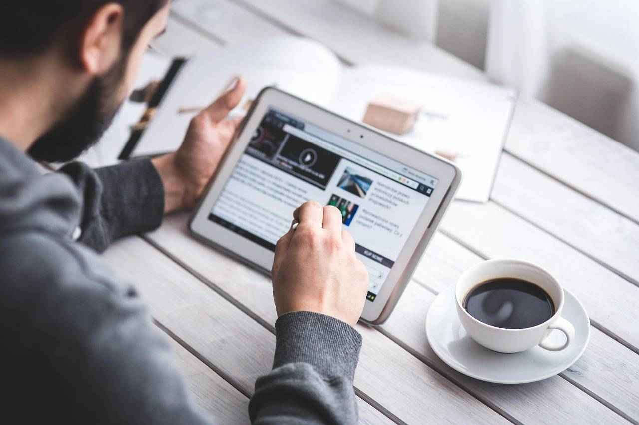 Un homme lit un article sur une tablette avec une tasse de café à ses côtés.
