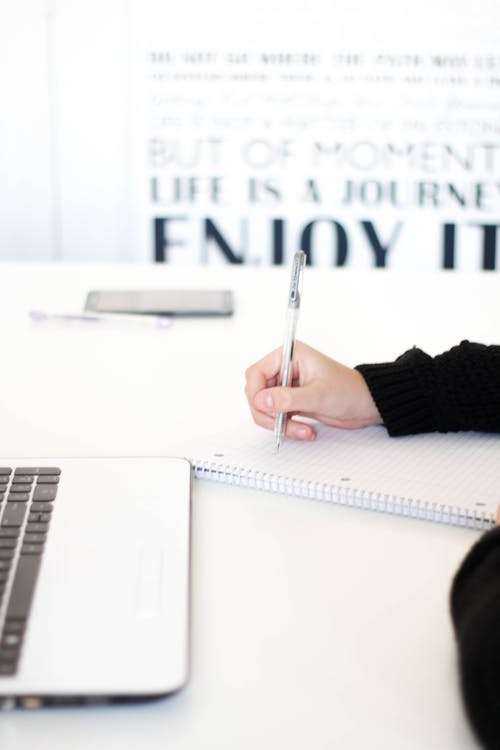 Personne prenant des notes sur un carnet, un ordinateur portable ouvert devant elle.