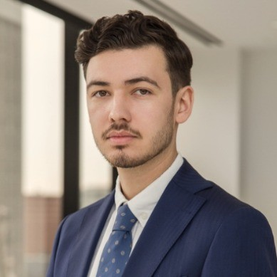 Portrait of a young man with short dark hair and trimmed beard wearing a navy blue suit, white shirt, and blue patterned tie in a modern office setting.