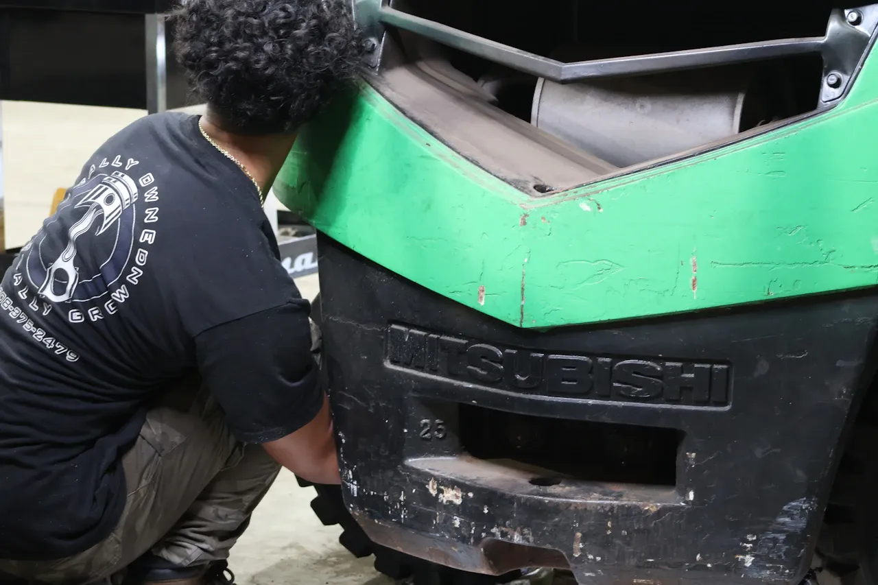 Heavy equipment repair on a Mitsubishi forklift as a technician inspects and services components beneath the engine cover.