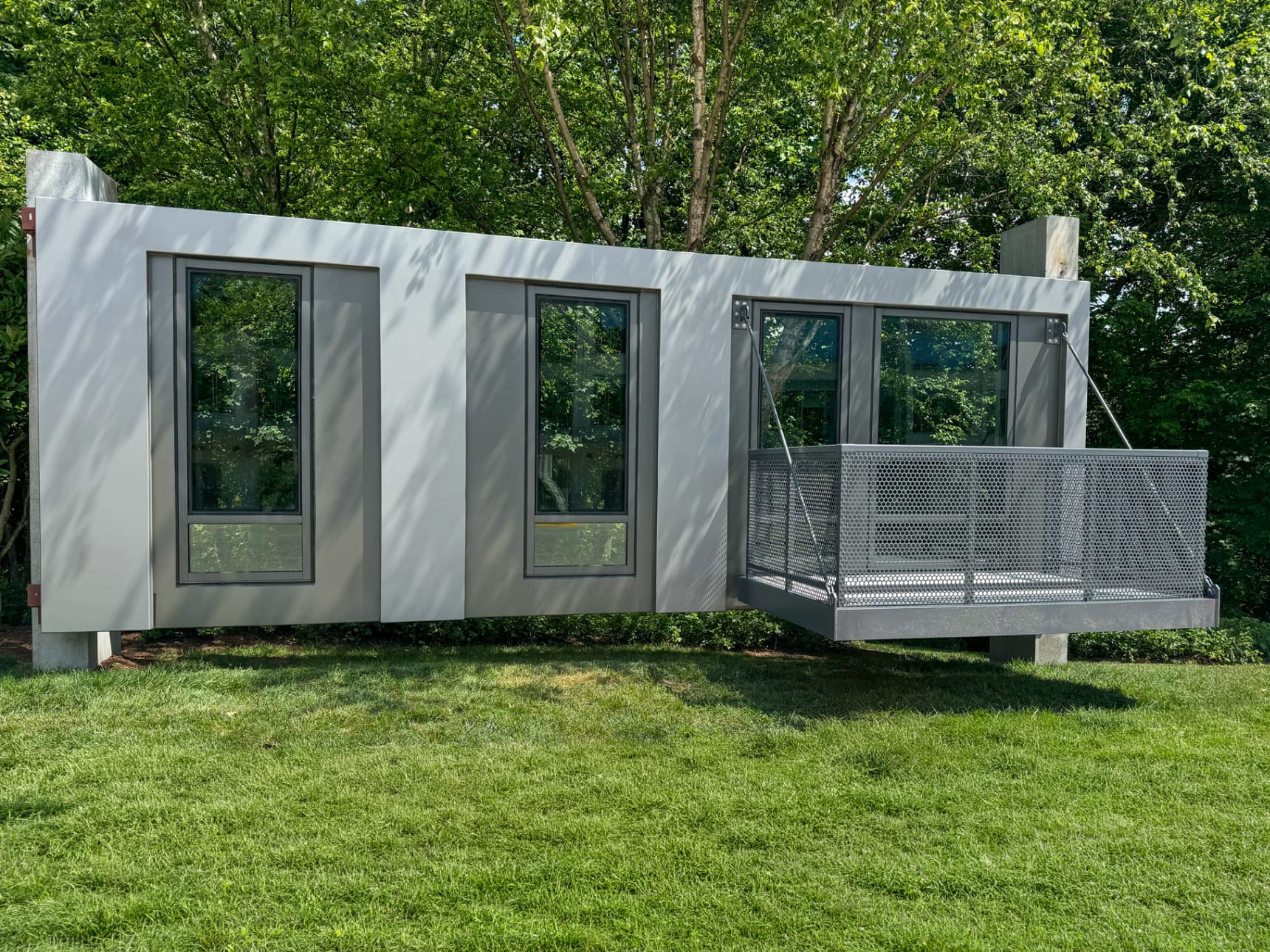 Modern modular housing unit with three tall windows and a suspended metal balcony over grass lawn.