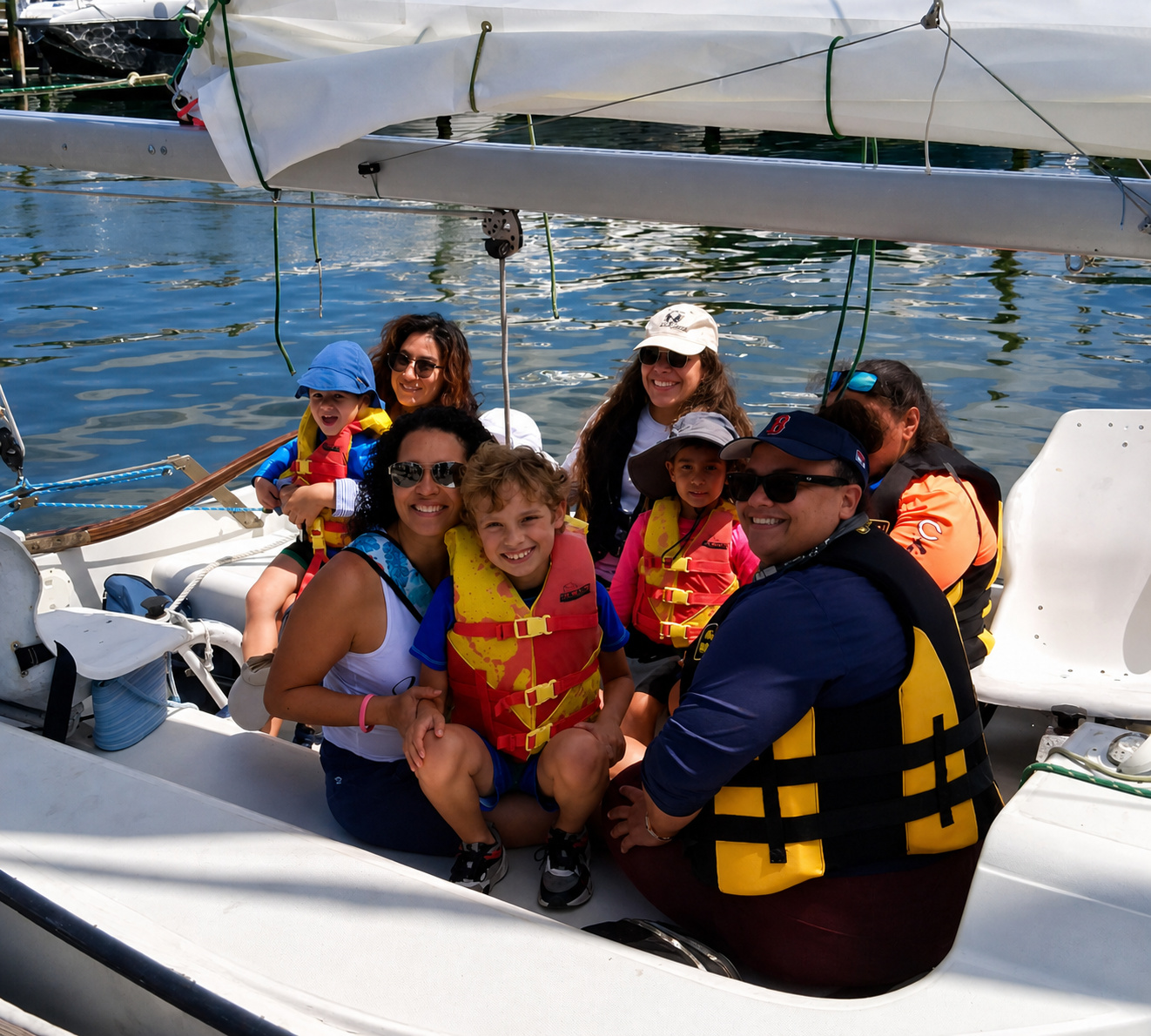 Children and adults in life vests sit together in the cockpit of a sailboat, smiling