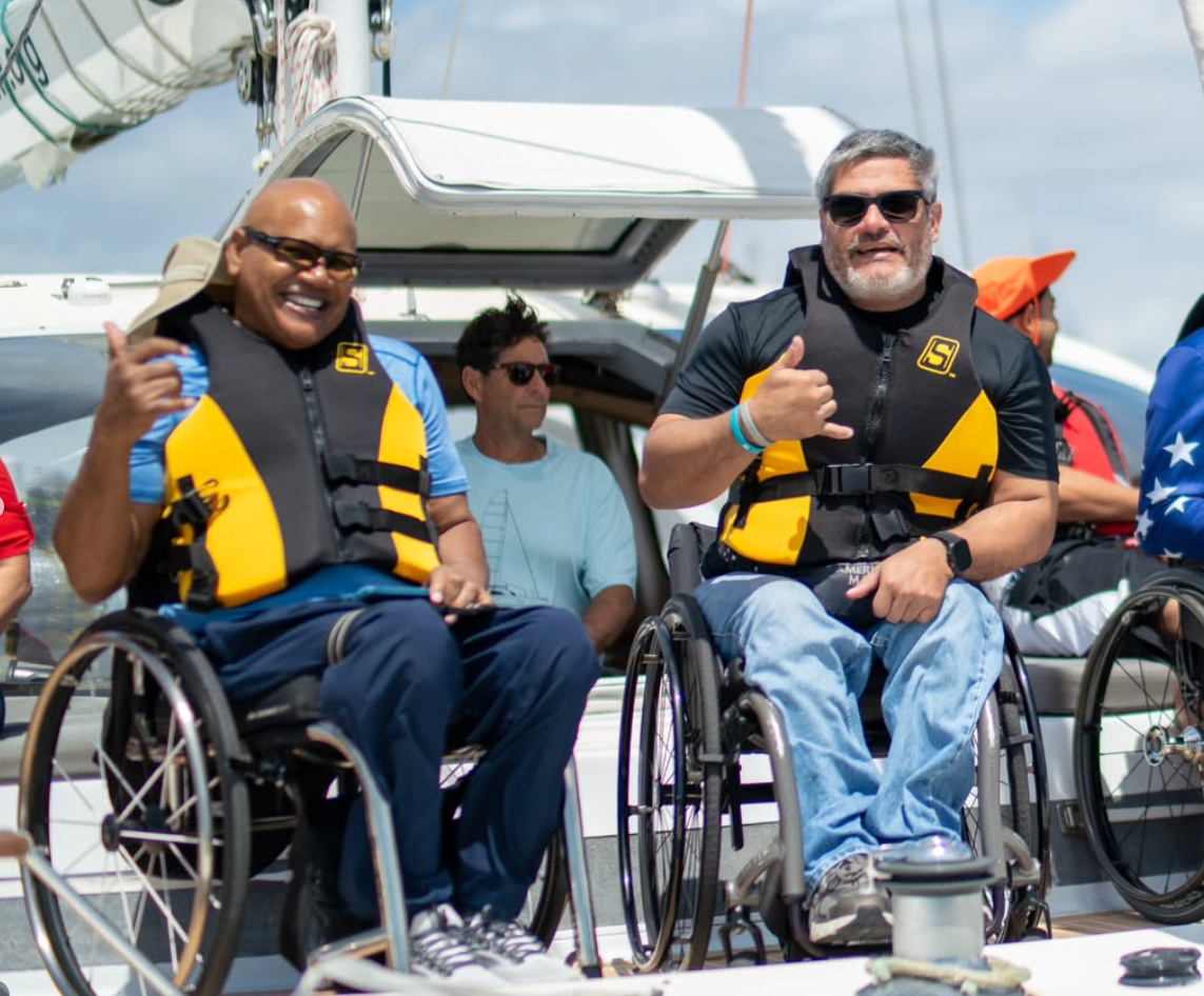 Two men who use wheelchairs, in yellow and black life vests, smile and make a hang-loose sign on the deck of a sailboat