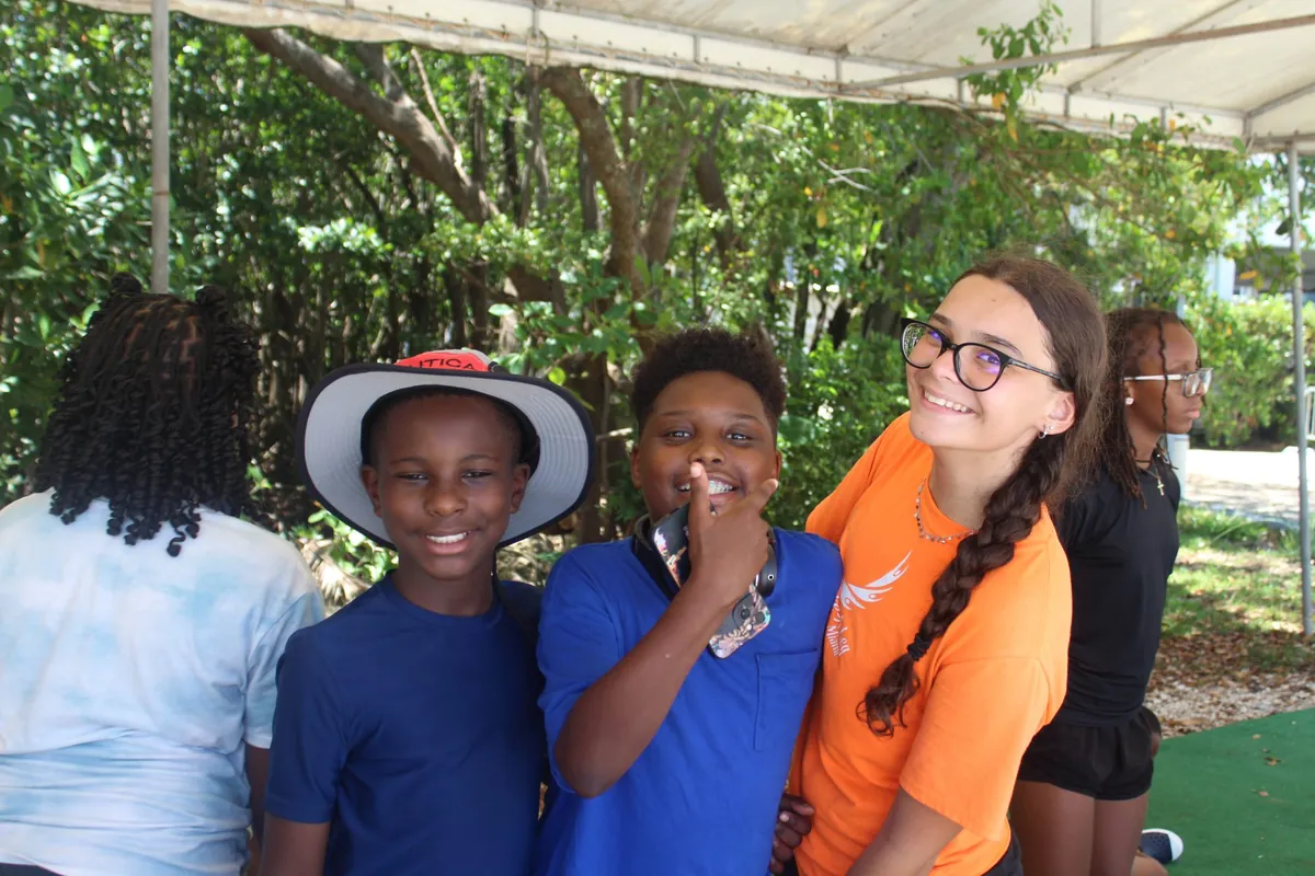 A teen mentor in an orange Shake-A-Leg shirt smiling with two boys under a shade tent
