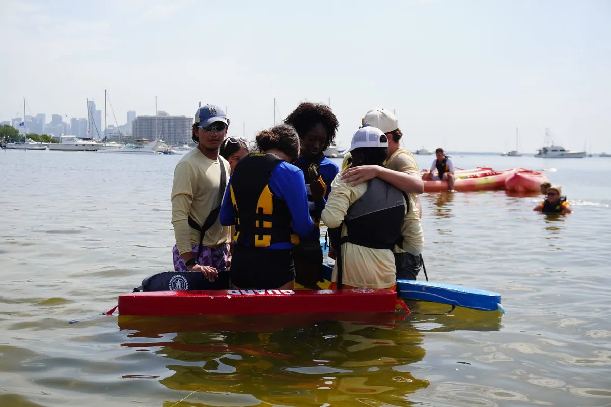 Outdoor educators and campers gathered around a rescue board in the shallows, with the Miami skyline behind