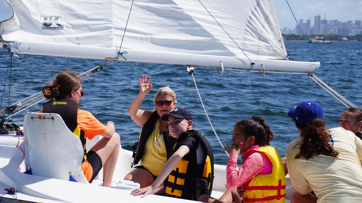 Campers and staff in life vests sail on Biscayne Bay, one waving, with the downtown Miami skyline behind them