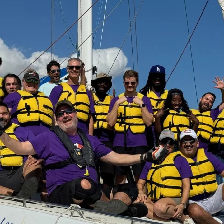 A company team in matching purple T-shirts and yellow life vests cheers aboard a catamaran at the dock