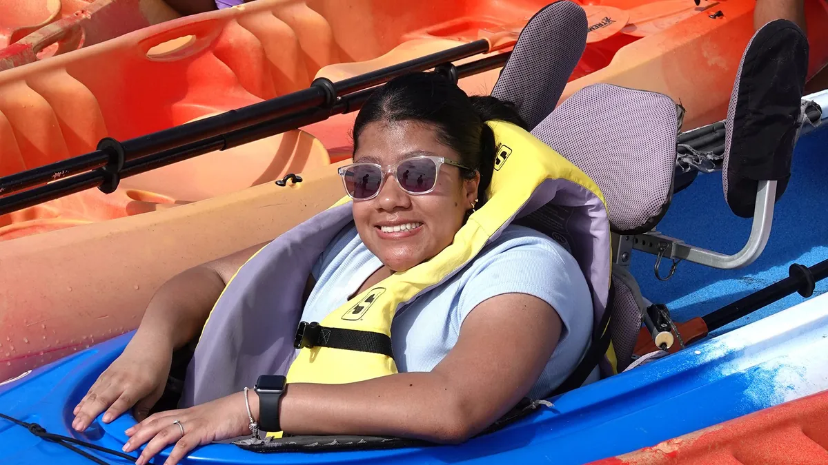 A smiling paddler in a yellow life vest sits in a blue kayak fitted with a padded high-back seat