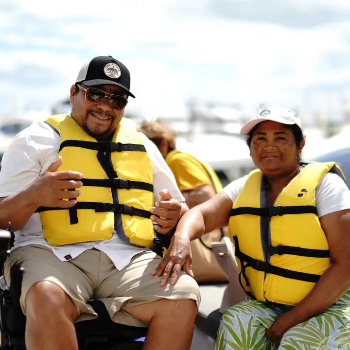 A smiling man in a wheelchair gives a thumbs up beside a smiling woman, both in yellow life vests aboard a boat