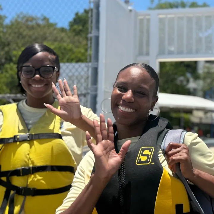 Two outdoor educators in life vests smile and wave by the dock gate