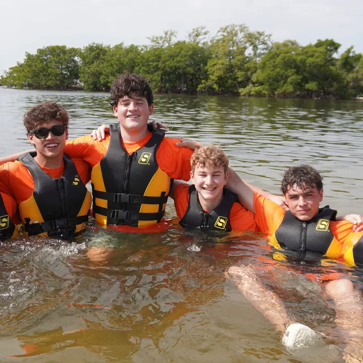 Teen mentors in orange shirts and life vests stand arm in arm, waist-deep in Biscayne Bay