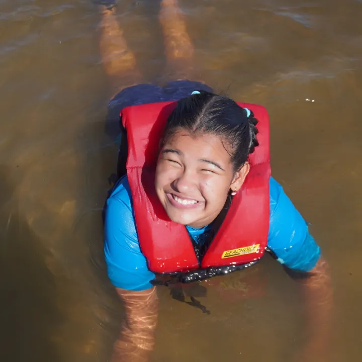 A camper in a red life jacket laughs while floating in the bay