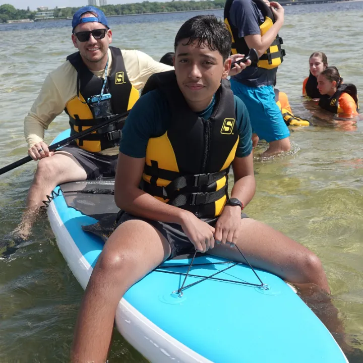 A camper sits on a paddleboard in the bay, with a counselor behind