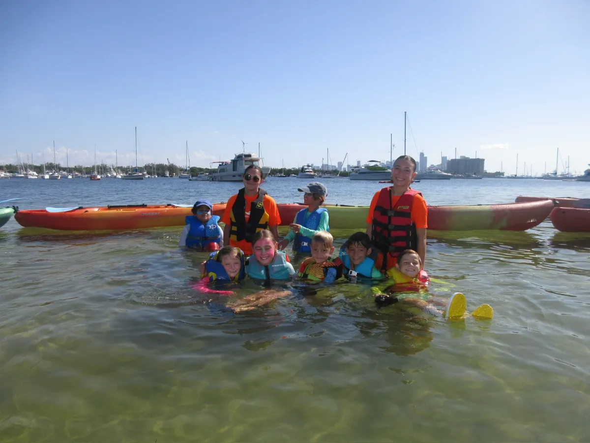 Campers and counselors in life jackets play in the island shallows next to their kayaks