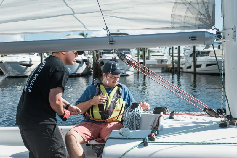 An instructor coaches a sailor in a life vest on the deck of a sailboat at the dock
