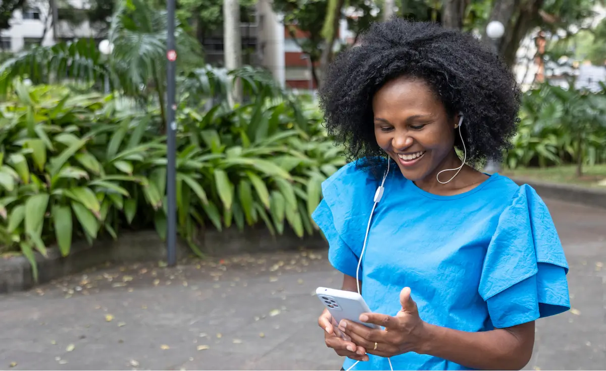 Smiling woman with curly hair in blue dress using phone and earphones outdoors by green plants.