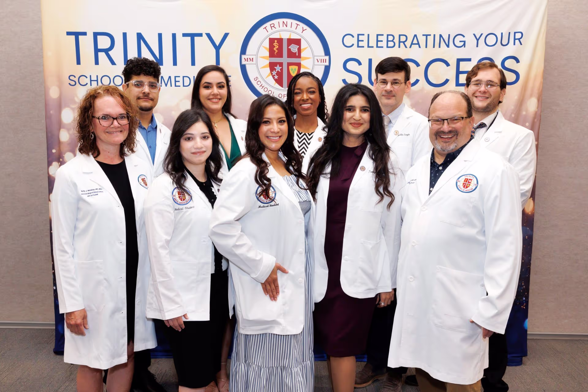 A group of medical students with both of the school Deans, standing in front of a Trinity Medical School sign during their white coat ceremony.
