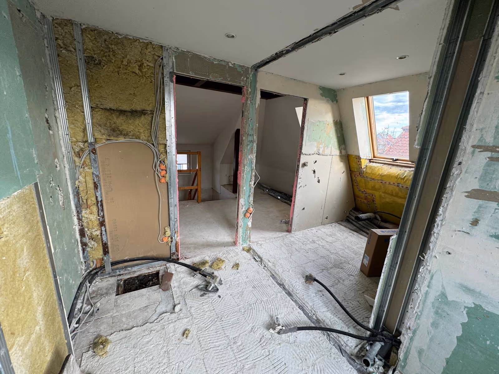 Wohnungsrenovierung in Stuttgart mit Teilsanierung im Dachgeschoss: Renovierungszustand einer Wohnung in Stuttgart mit sichtbarer Entkernung, offenliegenden Wandkonstruktionen, teilweise eingebauter Dämmung, vorbereiteten Elektro- und Sanitärleitungen, freiliegendem Estrichboden und unvollständigen Trockenbauflächen. Der Raum befindet sich im Umbau mit Blick auf Rohinstallation, Dämmvorbereitung, Raumaufteilung und anstehende Ausbauarbeiten.
