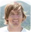 Young person with light brown hair smiling outdoors near water and greenery.