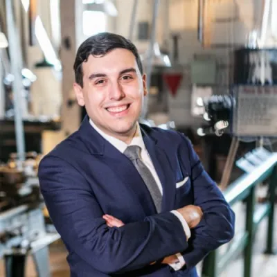 Young man in a navy suit and tie smiling with arms crossed in a modern office setting.