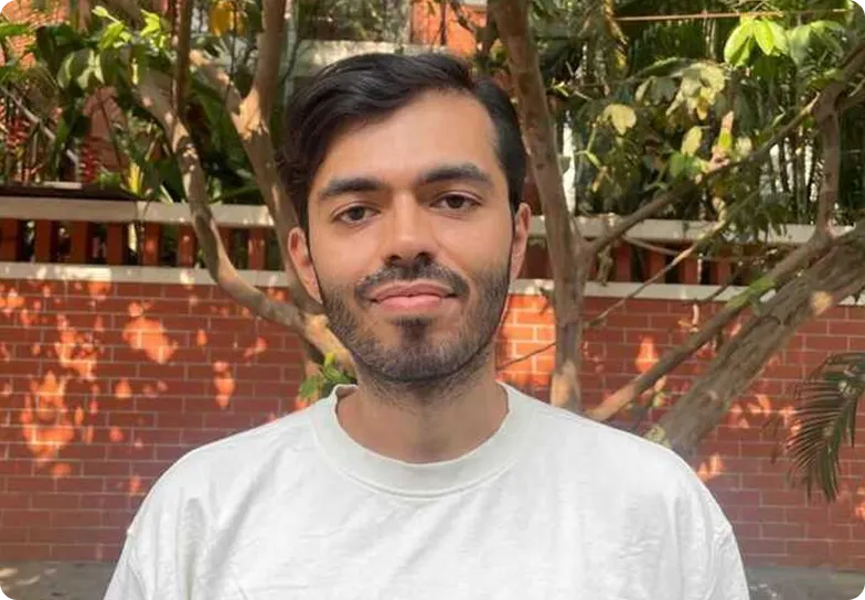 Man with short dark hair and beard wearing a white shirt standing outdoors by a brick wall and trees.