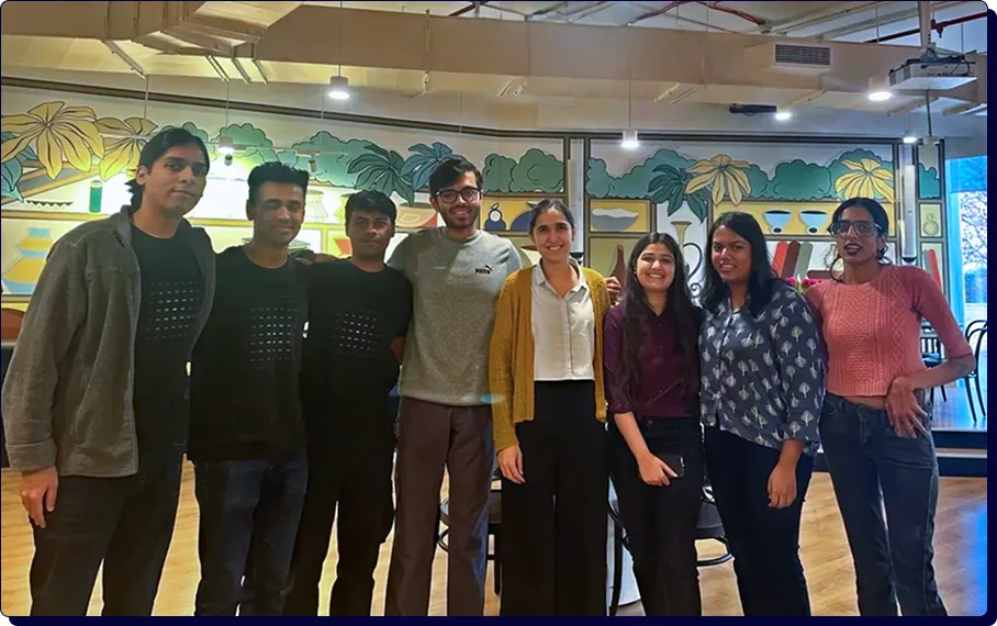 Group of eight young adults standing indoors smiling, against a colorful wall mural with plants and vases.