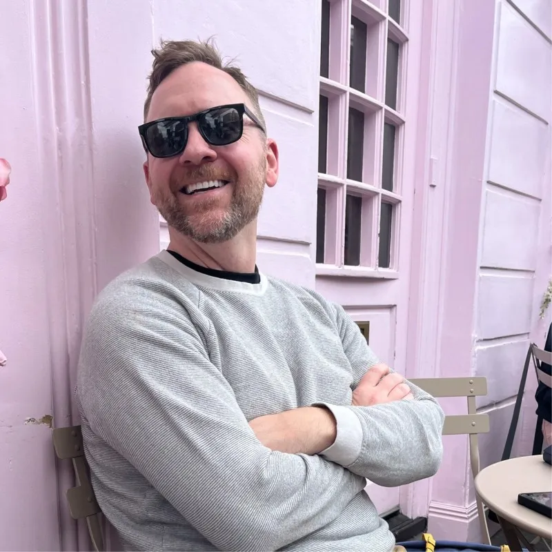 Smiling man with sunglasses and beard wearing a gray sweater, sitting with arms crossed by a pastel pink wall.