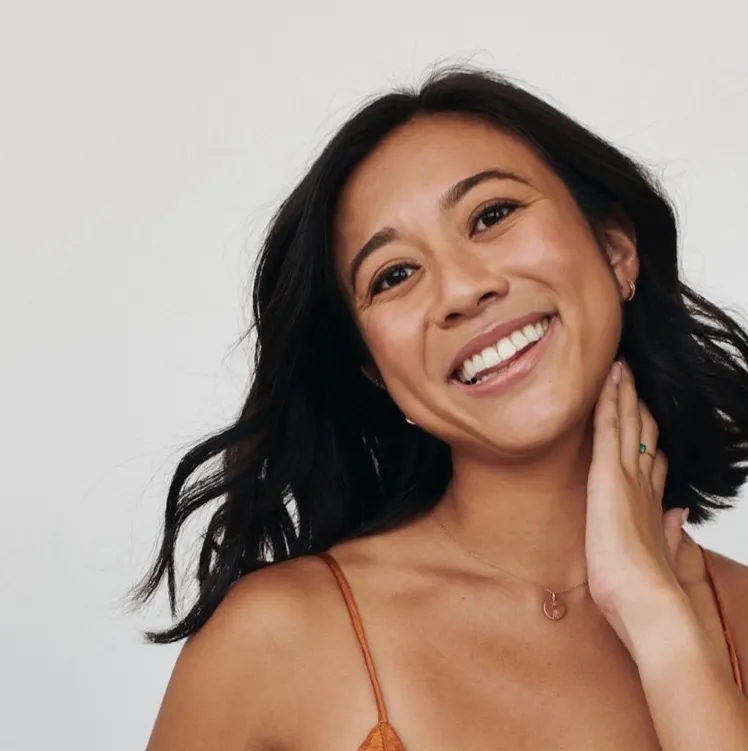 Smiling woman with dark hair wearing a thin strap top and a pendant necklace against a plain background.