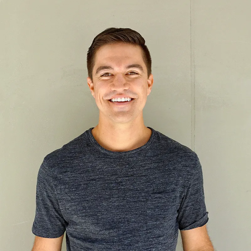 Smiling young man with short dark hair wearing a dark gray t-shirt against a plain wall.