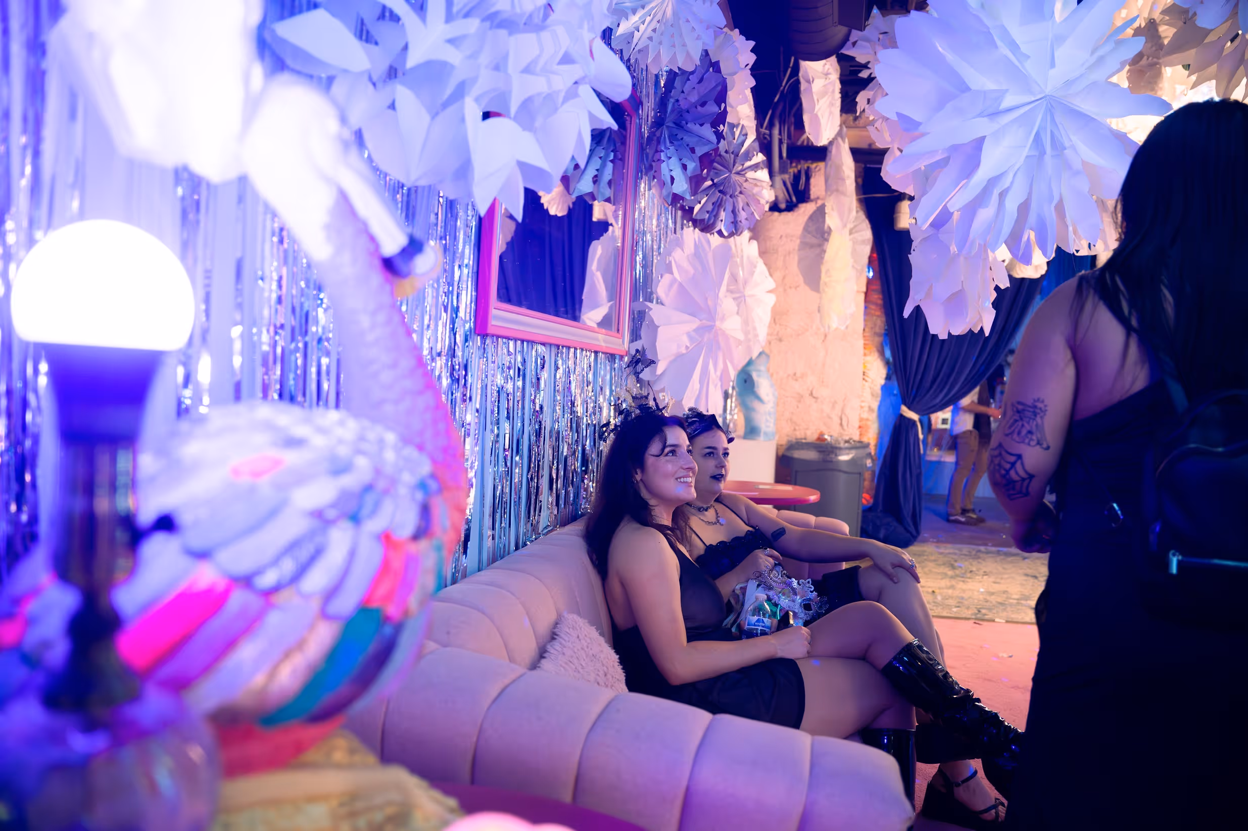 Seated guests in a softly lit lounge area, resting on a blush-colored sofa beneath white paper-flower decorations and pastel lighting, engaged in conversation against a backdrop of sheer curtains and ambient glow.