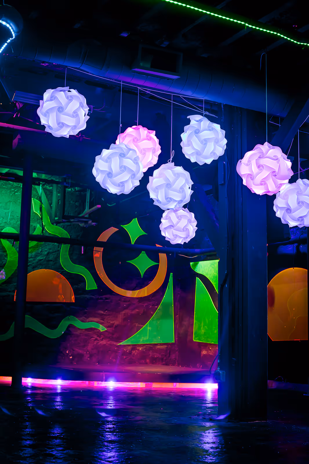 Neon-lit event space with hanging white geometric pendant lights, colorful abstract wall art, and an open polished concrete floor in an industrial venue.