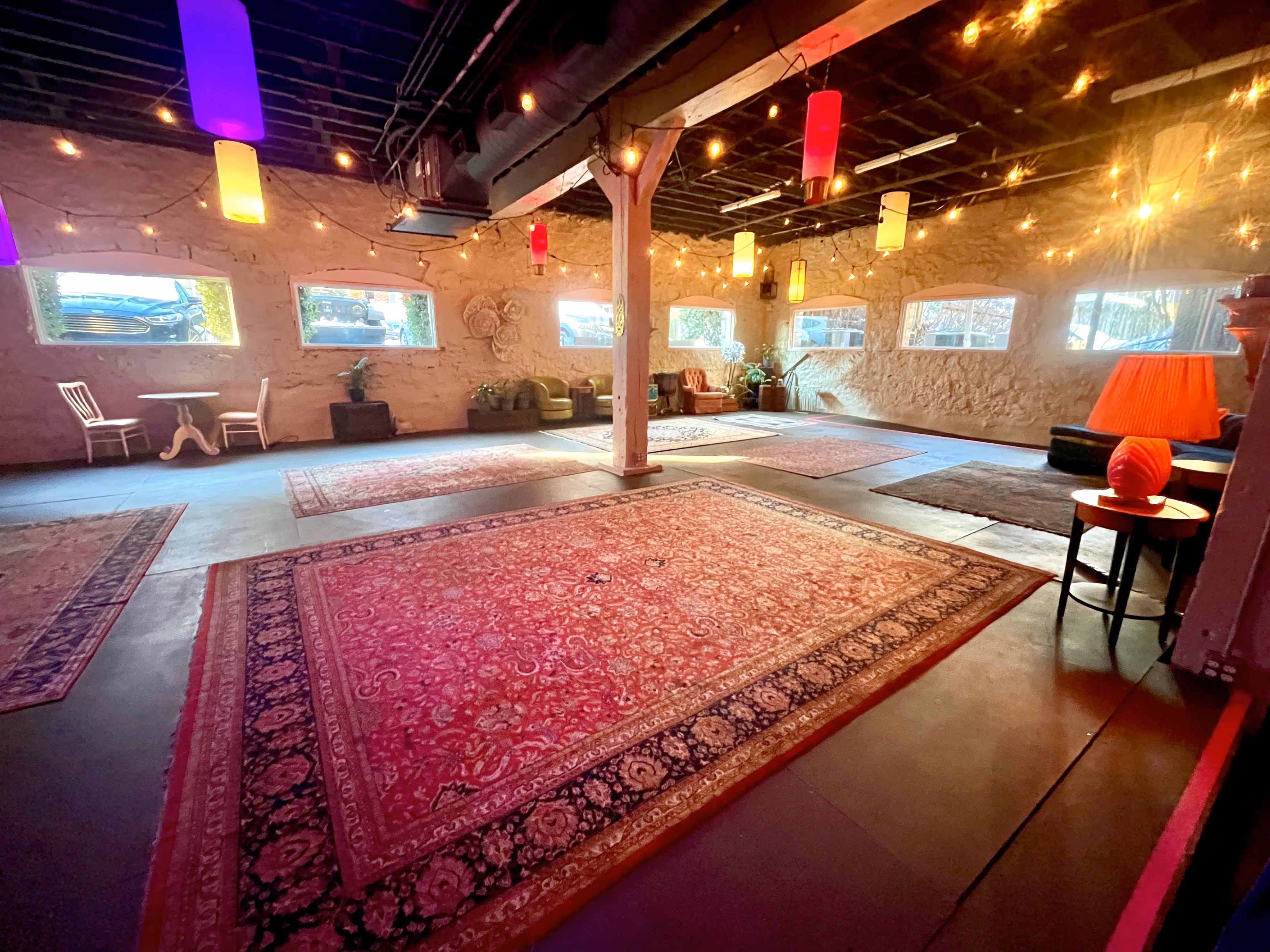 A spacious event hall with exposed ceiling beams, string lights and multicolored hanging fixtures, a large ornate rug on the floor, lounge seating along the walls, and warm ambient light spilling in through windows.