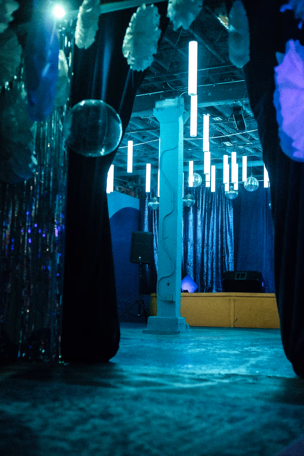 Blue Room stage setup with shimmering blue curtains, hanging tube lights, disco balls, and a speaker, viewed from behind dark drapes with a glowing, atmospheric ambiance.