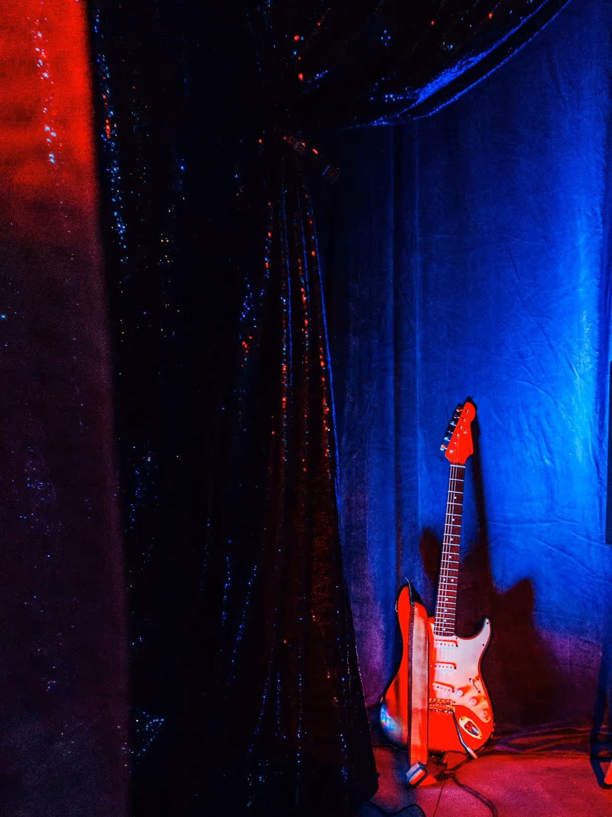 Blue Room with a red electric guitar resting against blue drapes beside a black sequined curtain, lit by moody red and blue stage lighting.