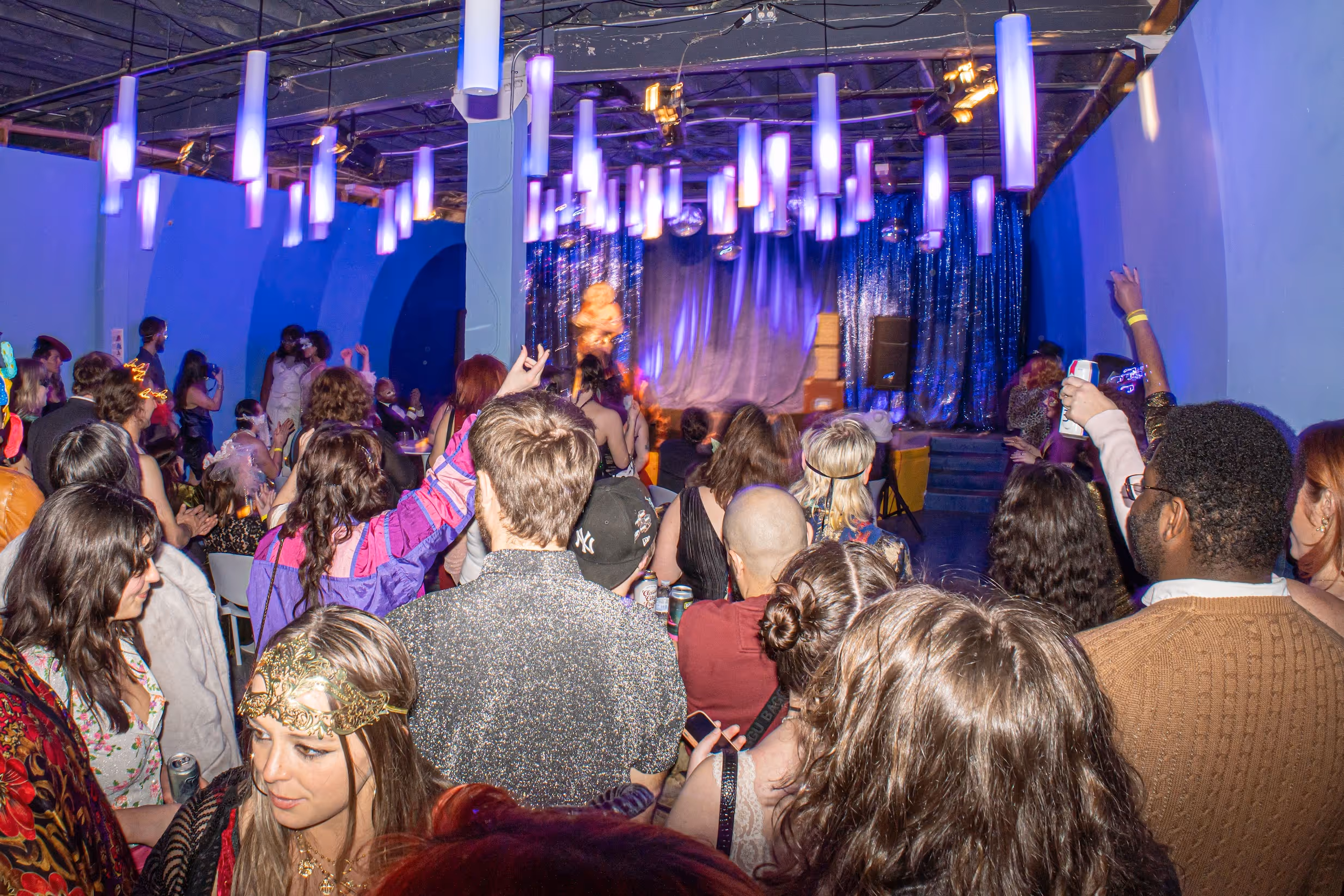 Blue Room filled with a lively crowd watching a performance on stage, featuring shimmering blue curtains, hanging tube lights glowing purple, and a festive atmosphere.