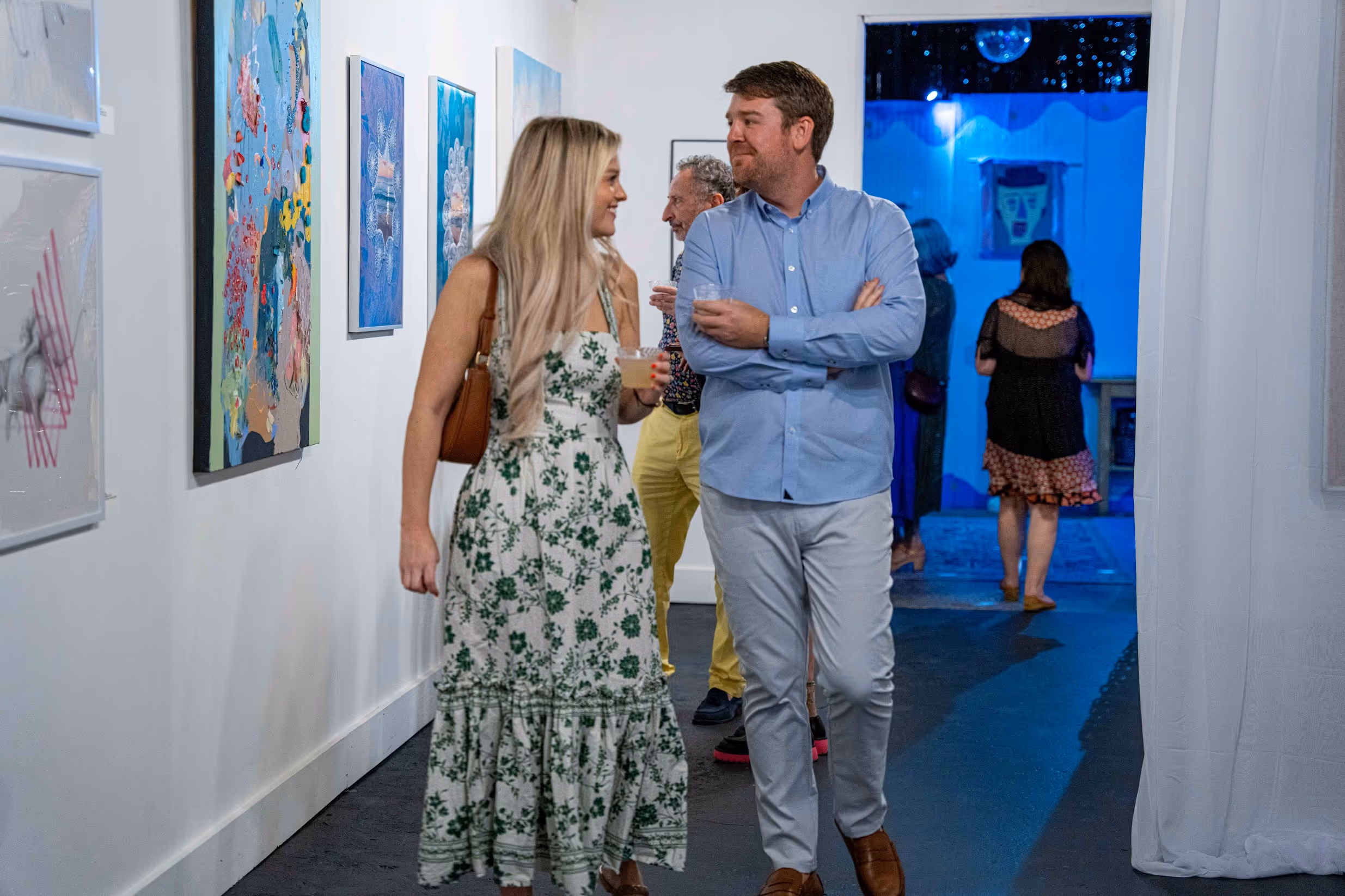 Art Gallery with visitors walking through a bright white-walled hallway lined with colorful contemporary paintings, leading into a blue-lit adjoining room where more artwork is displayed.